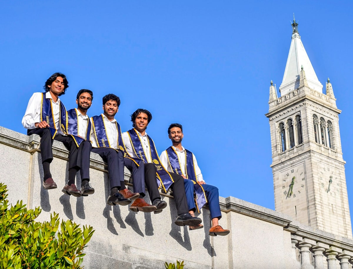 Five graduates in purple and gold stoles sit on a stone wall beside Sather Tower at UC Berkeley.