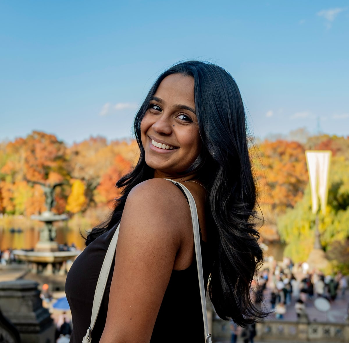 A woman with long dark hair smiles over her shoulder in Central Park, autumn trees behind her.