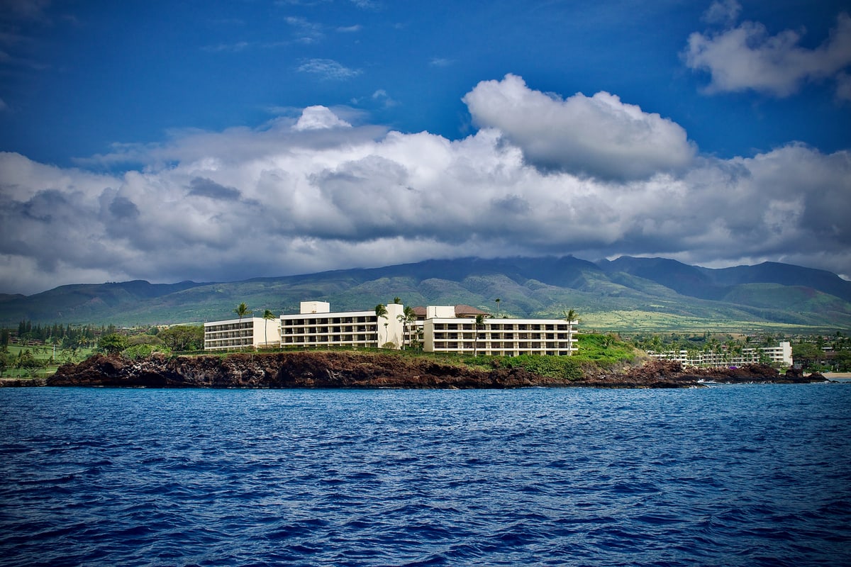 A white hotel sits on a green point beneath mountains and towering clouds.