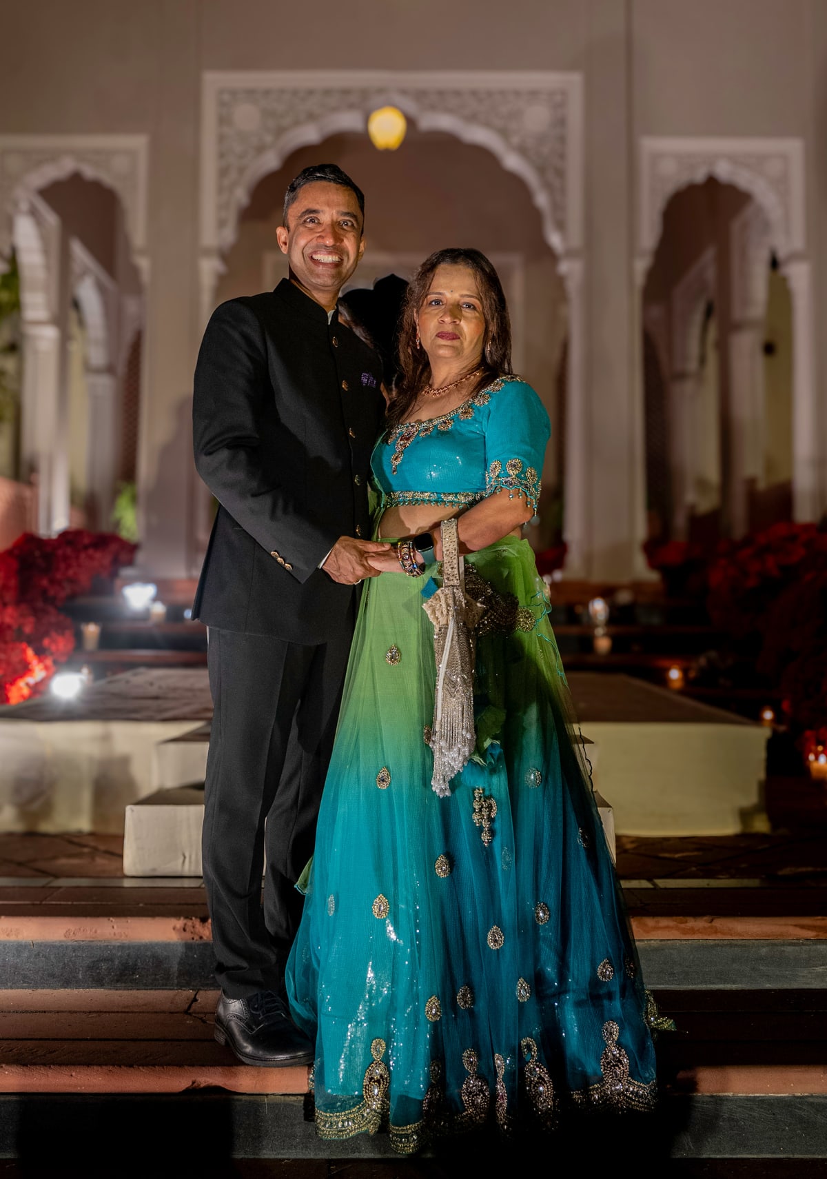 A couple in formal Indian attire poses beneath carved stone arches at dusk.