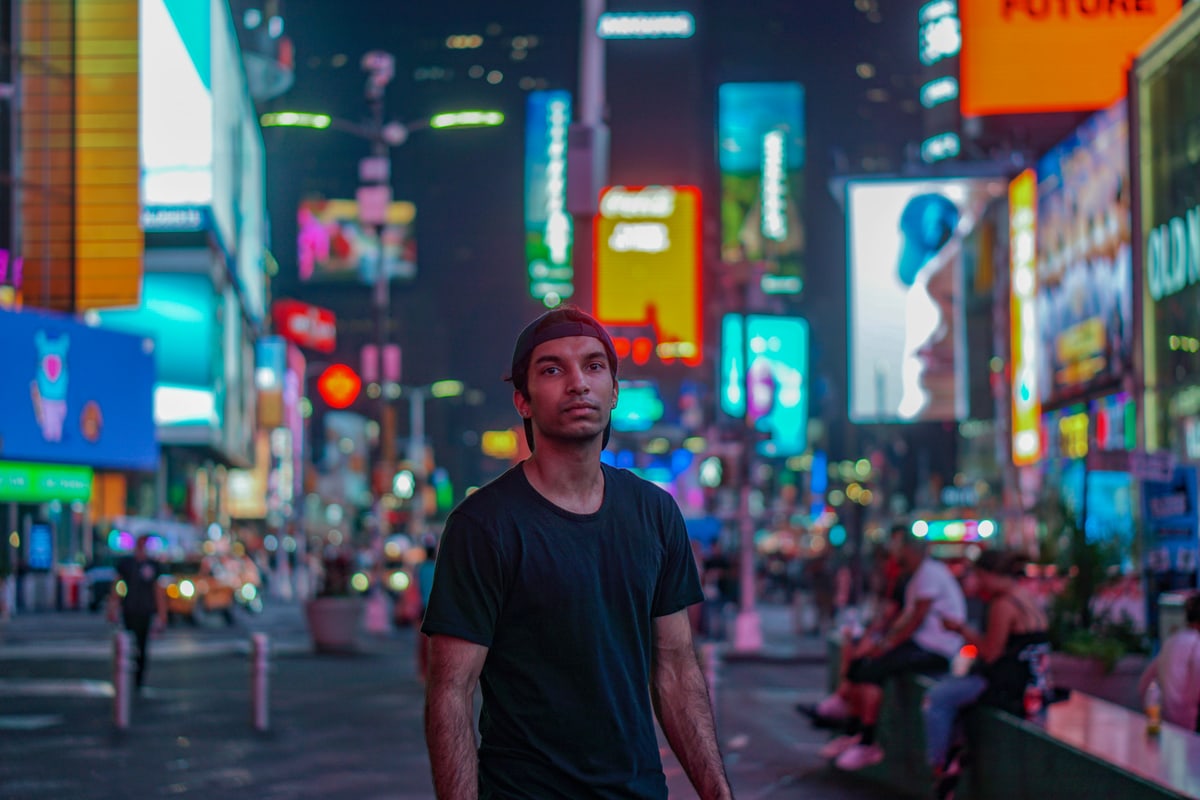 A man in a black T-shirt stands in Times Square at night, neon signs glowing behind him.