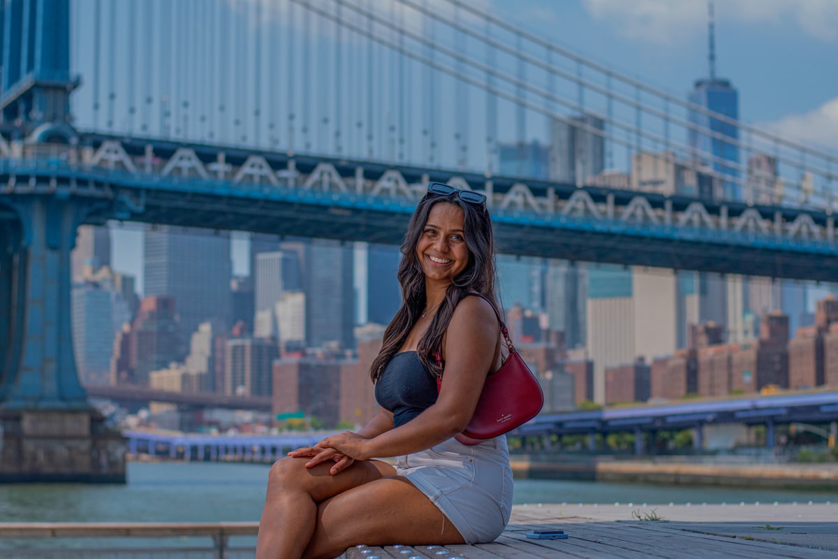 A woman sits on a waterfront ledge beneath the Manhattan Bridge with the city skyline behind her.