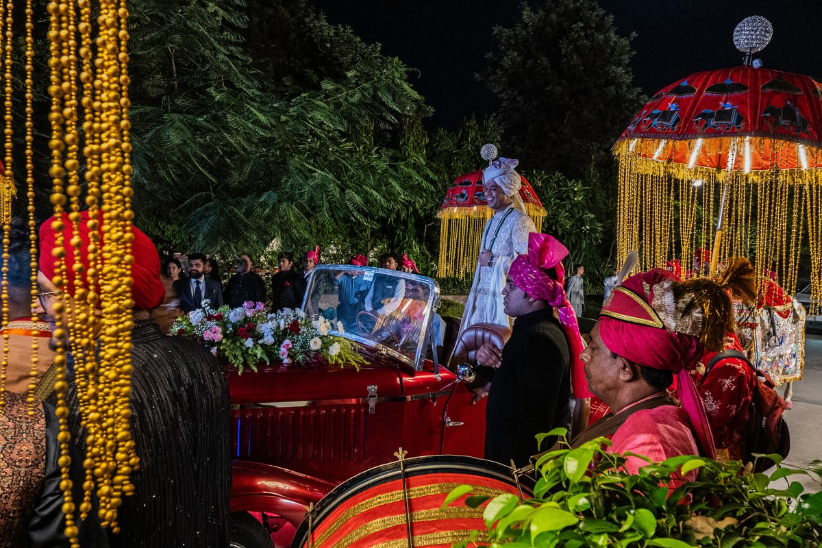 A groom rides a flower-covered vintage car through a lively baraat procession at night.