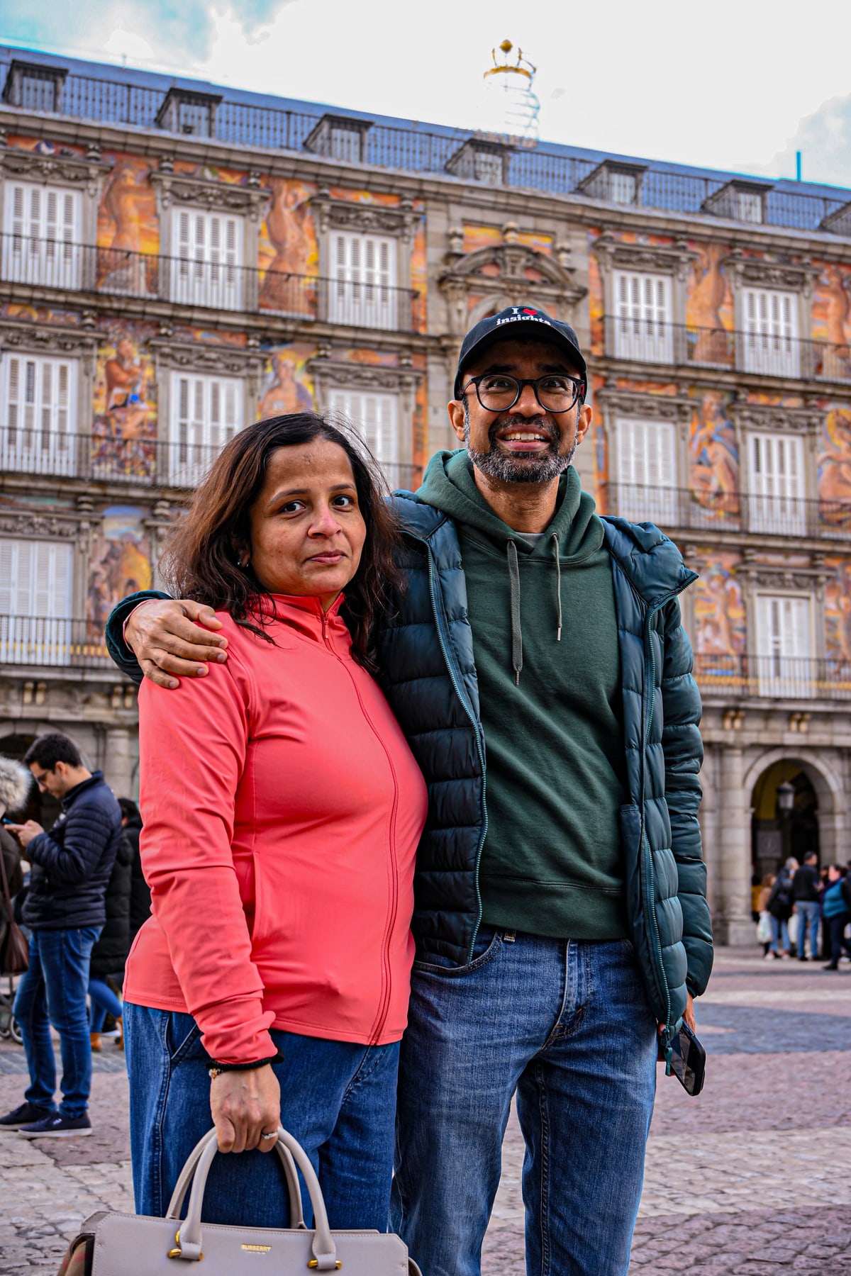 A smiling couple poses arm in arm in Plaza Mayor, painted facades behind them.
