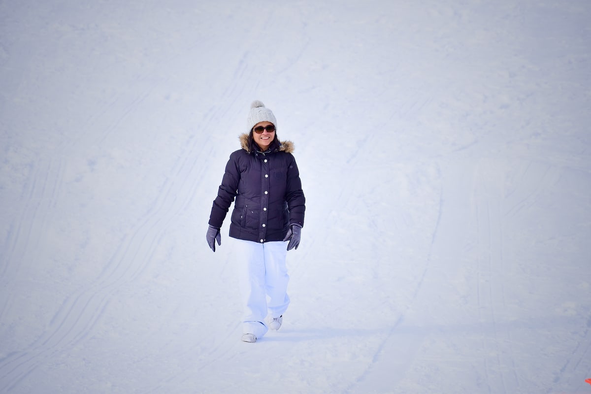 A woman in a puffy jacket and sunglasses walks across bright white snow.