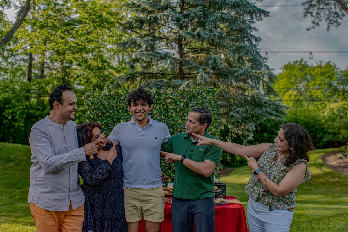 A graduate laughs with family members in a leafy backyard while his mother points at him.