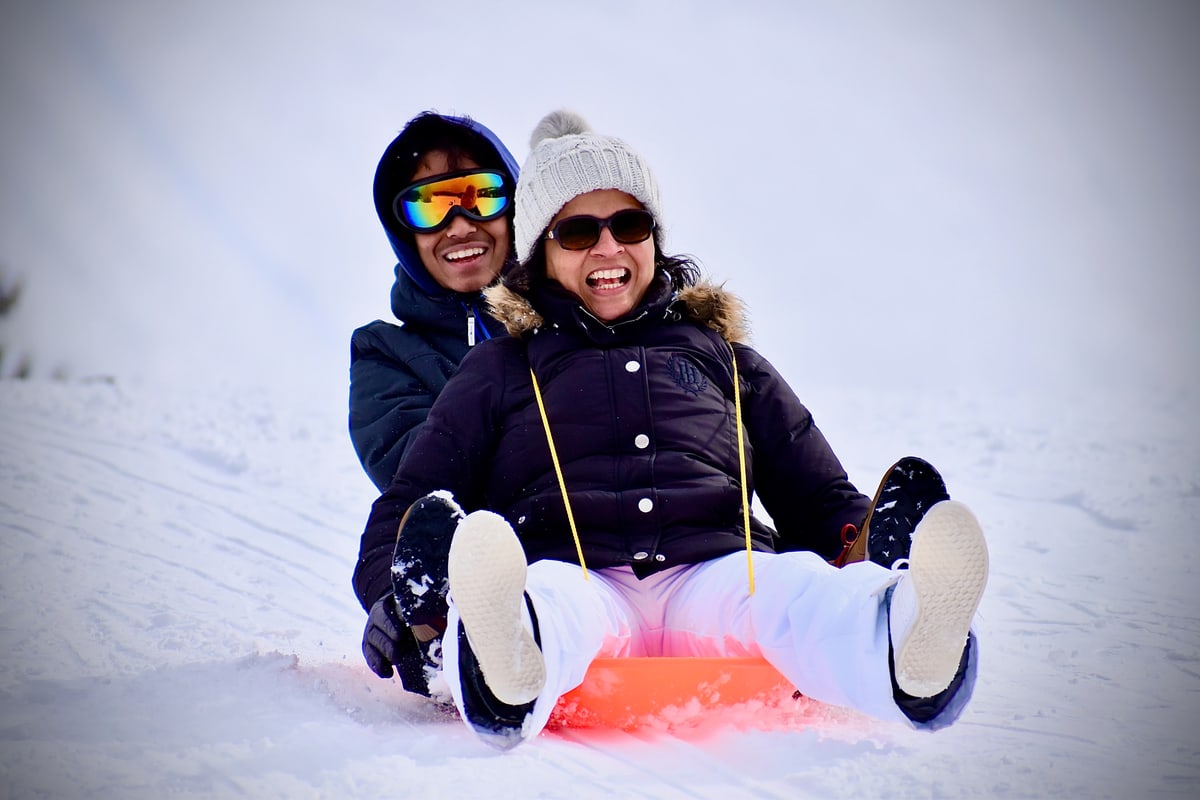 Two people laugh as they slide down a snowy hill on an orange sled.