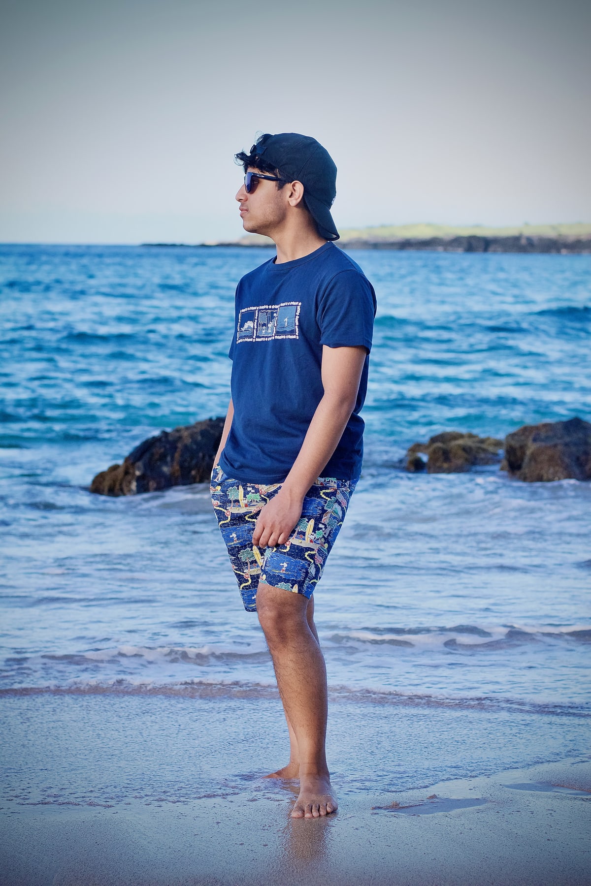 A young man in a cap and sunglasses stands barefoot in the surf, looking out to sea.