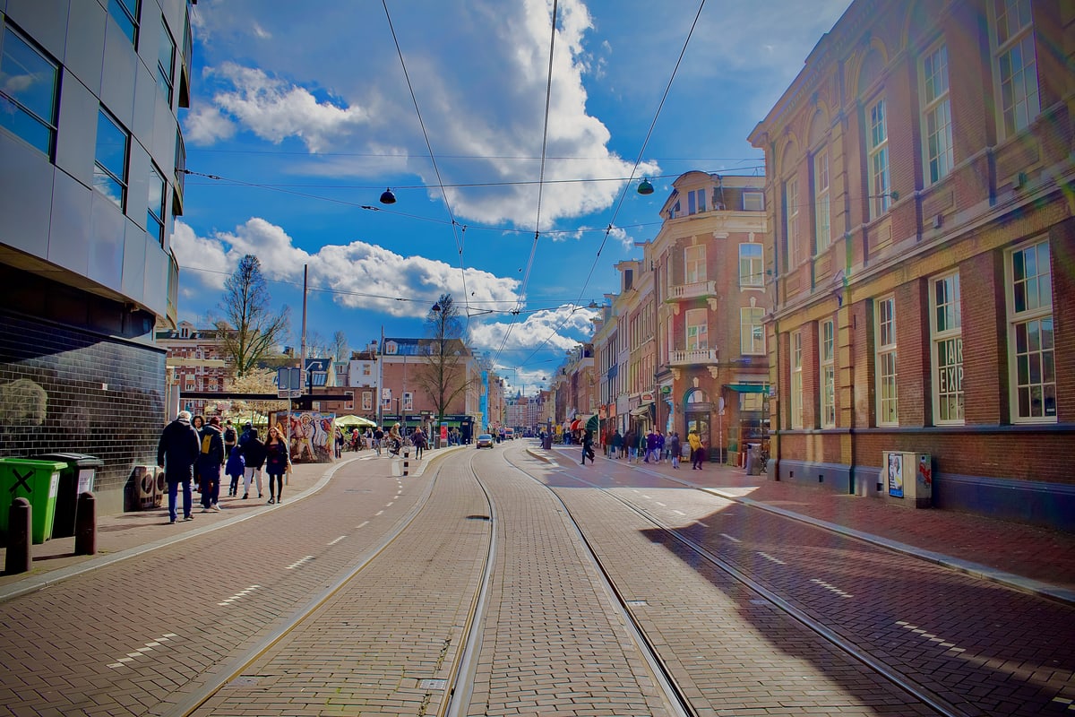 Tram tracks lead down a sunny Amsterdam street under puffy clouds.