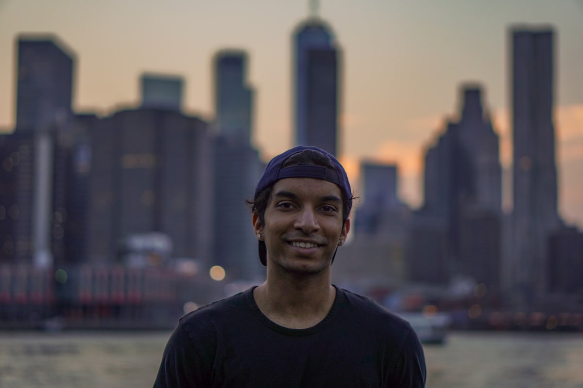 A young man in a black cap smiles in front of a softly blurred Manhattan skyline at dusk.