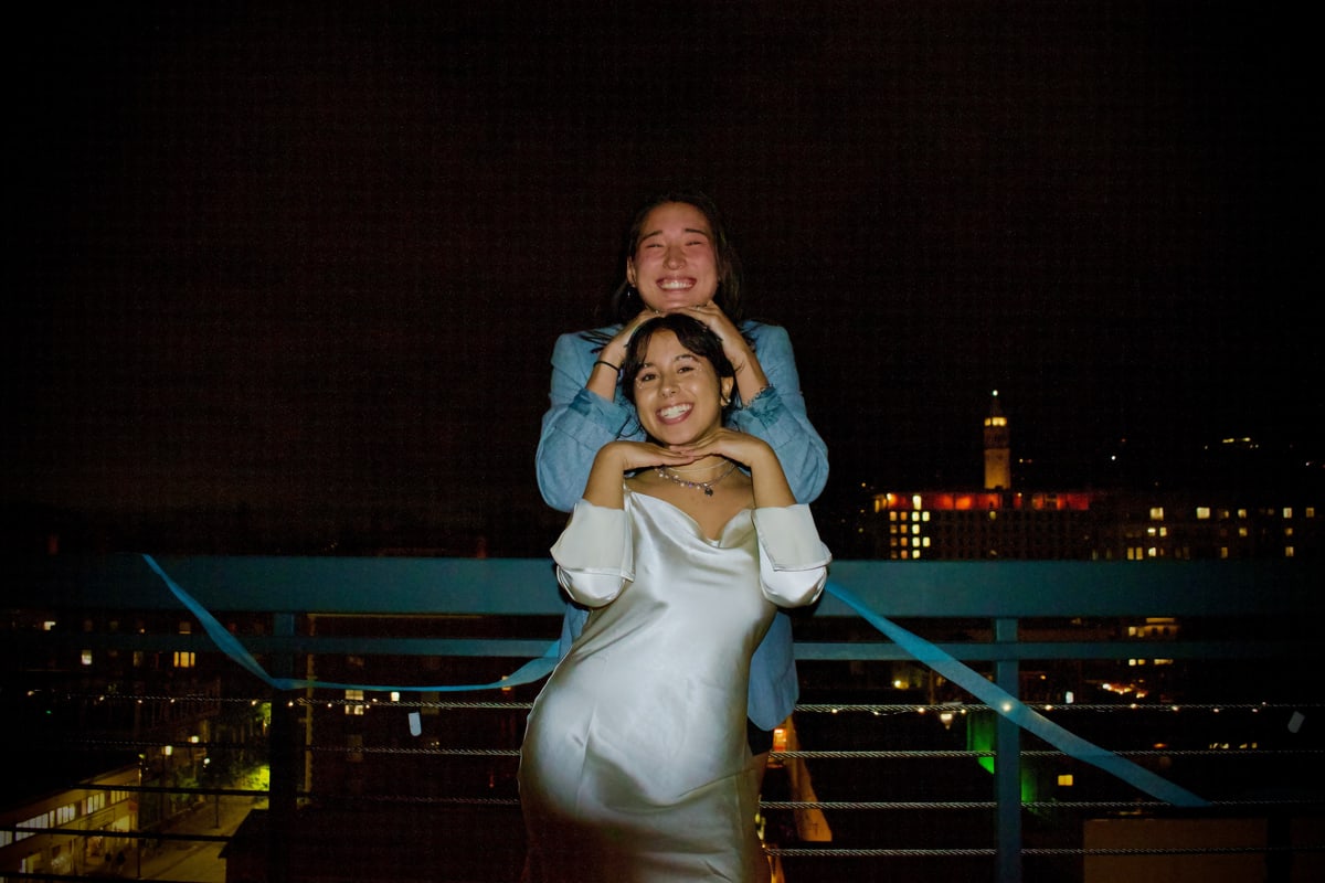 A woman in a white satin dress poses as a friend rests his hands on her head, the city lit up behind them.