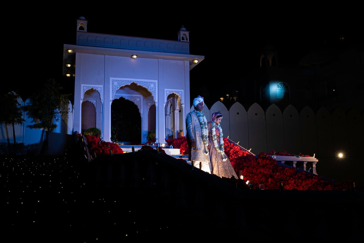 A bride and groom stand on lit palace steps beneath a white archway at night.