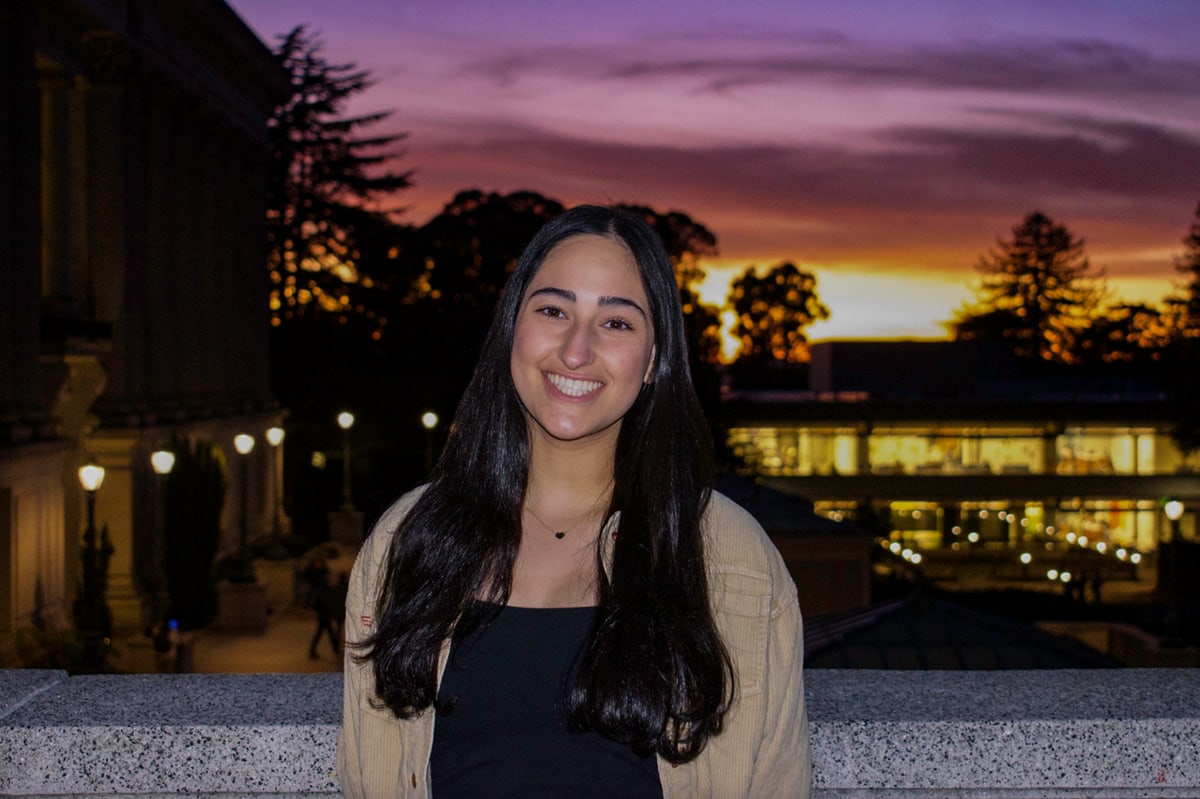 A woman with long dark hair smiles at sunset, the sky glowing orange behind a campus building.