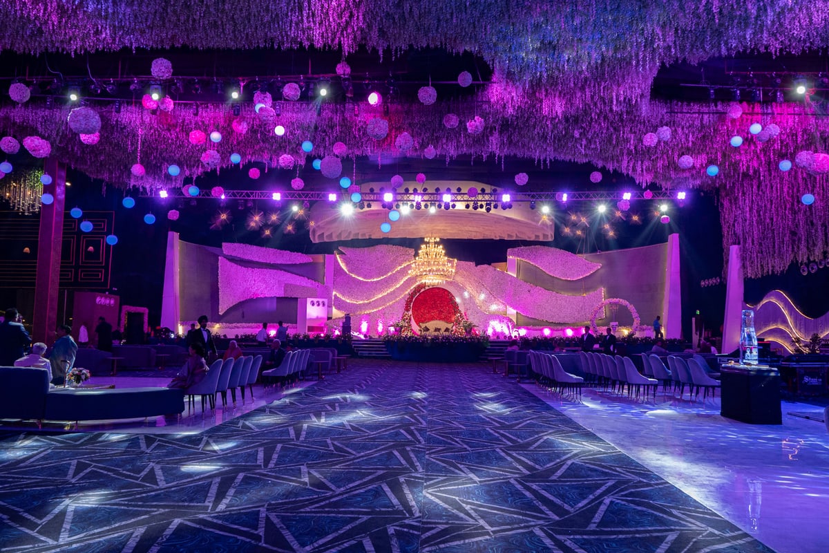 A vast reception hall glows purple beneath a ceiling of hanging flowers and chandeliers.