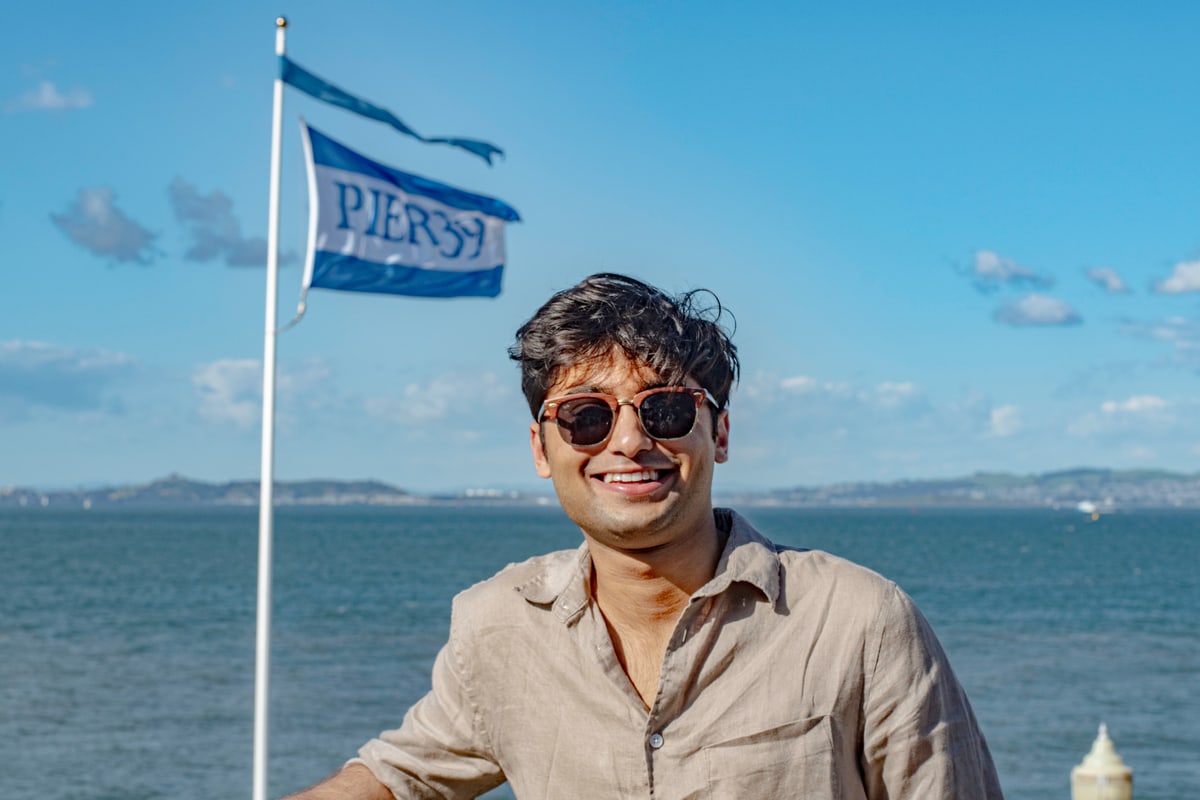 A man in sunglasses grins by the water beside a waving flag.