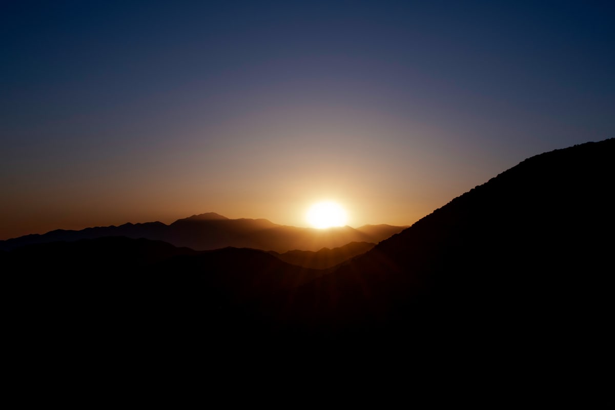 The sun flares over a dark mountain ridge at dusk.
