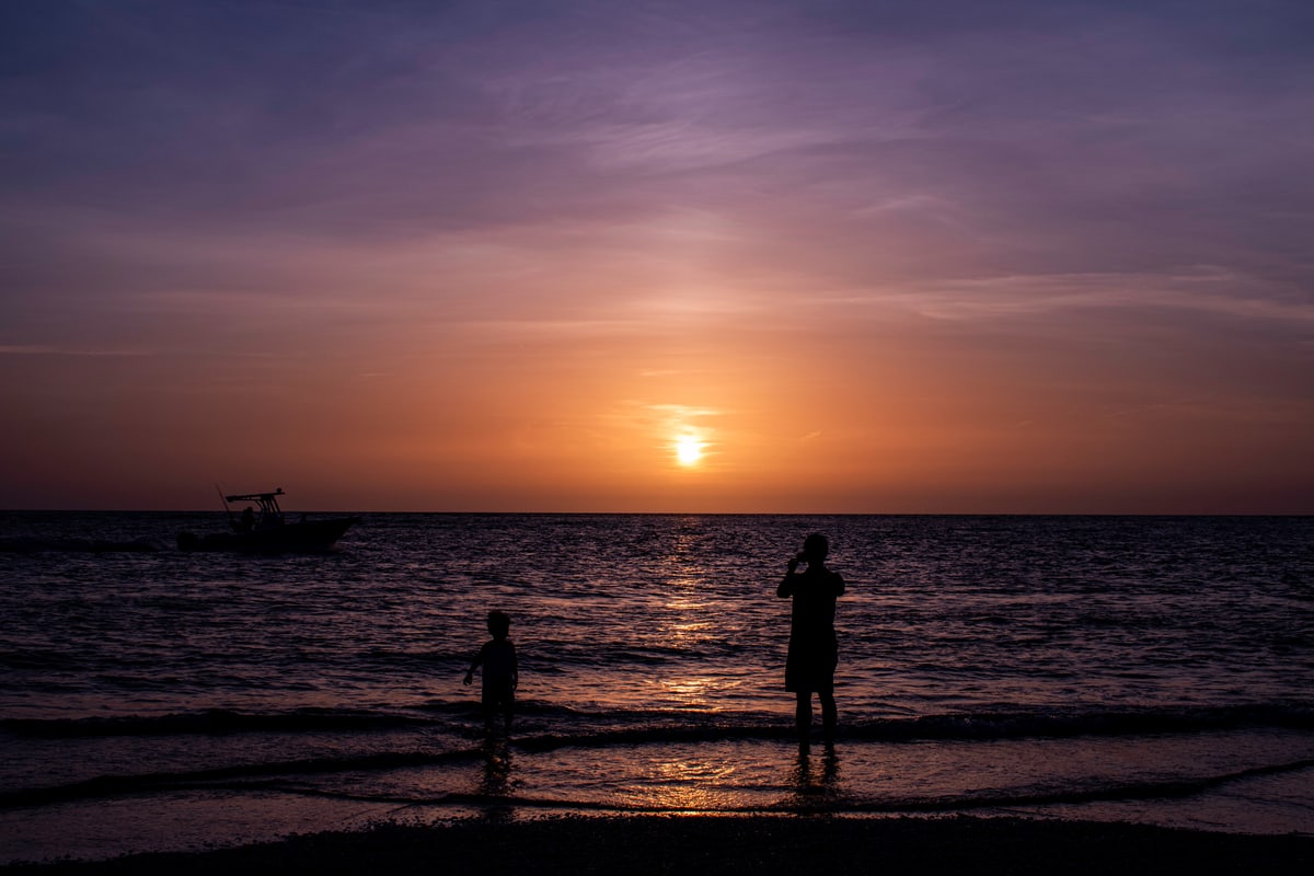 Two silhouettes stand in the shallows as the sun sets over the ocean.