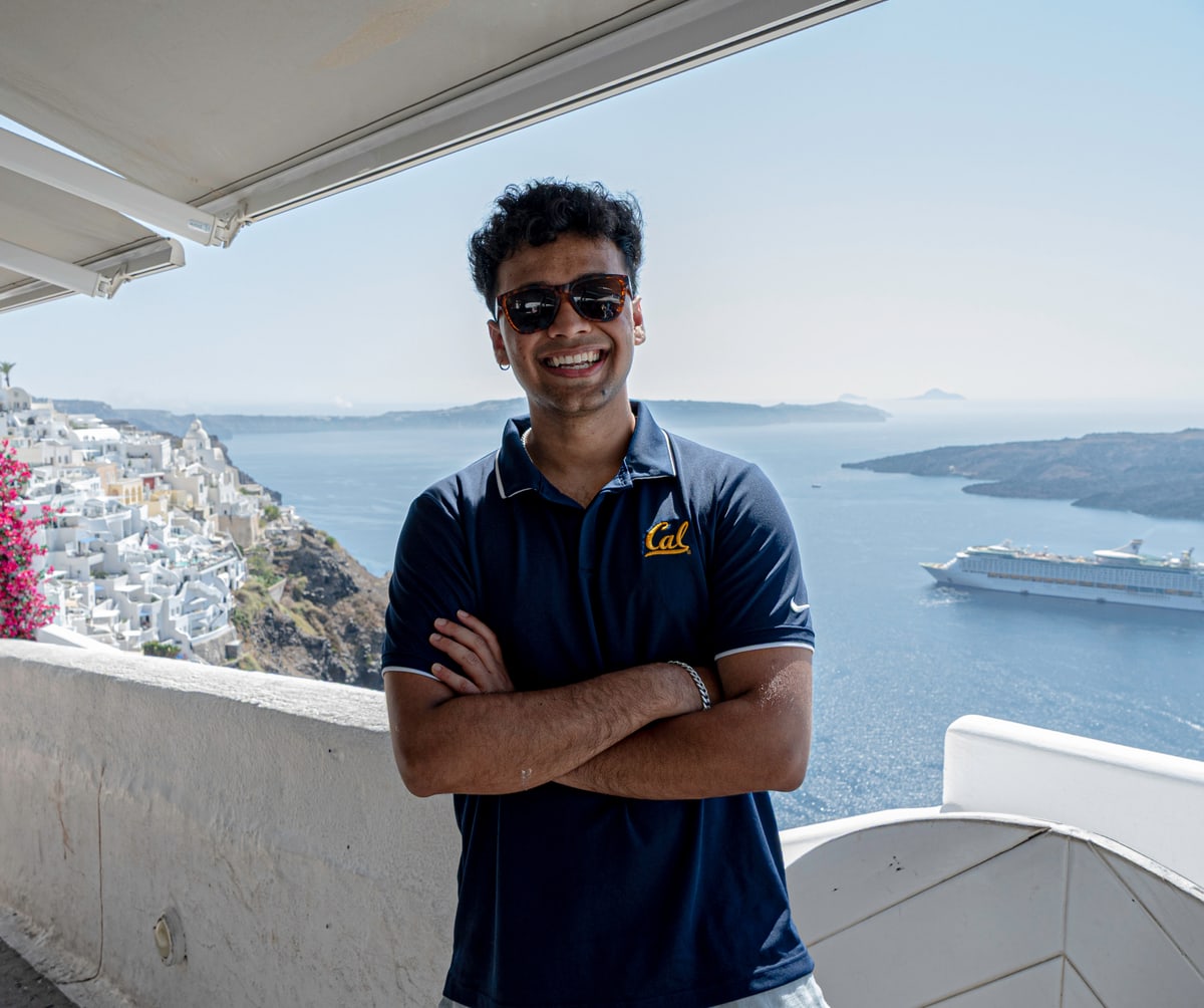 A man in sunglasses stands with arms crossed on a Santorini terrace above the sea.