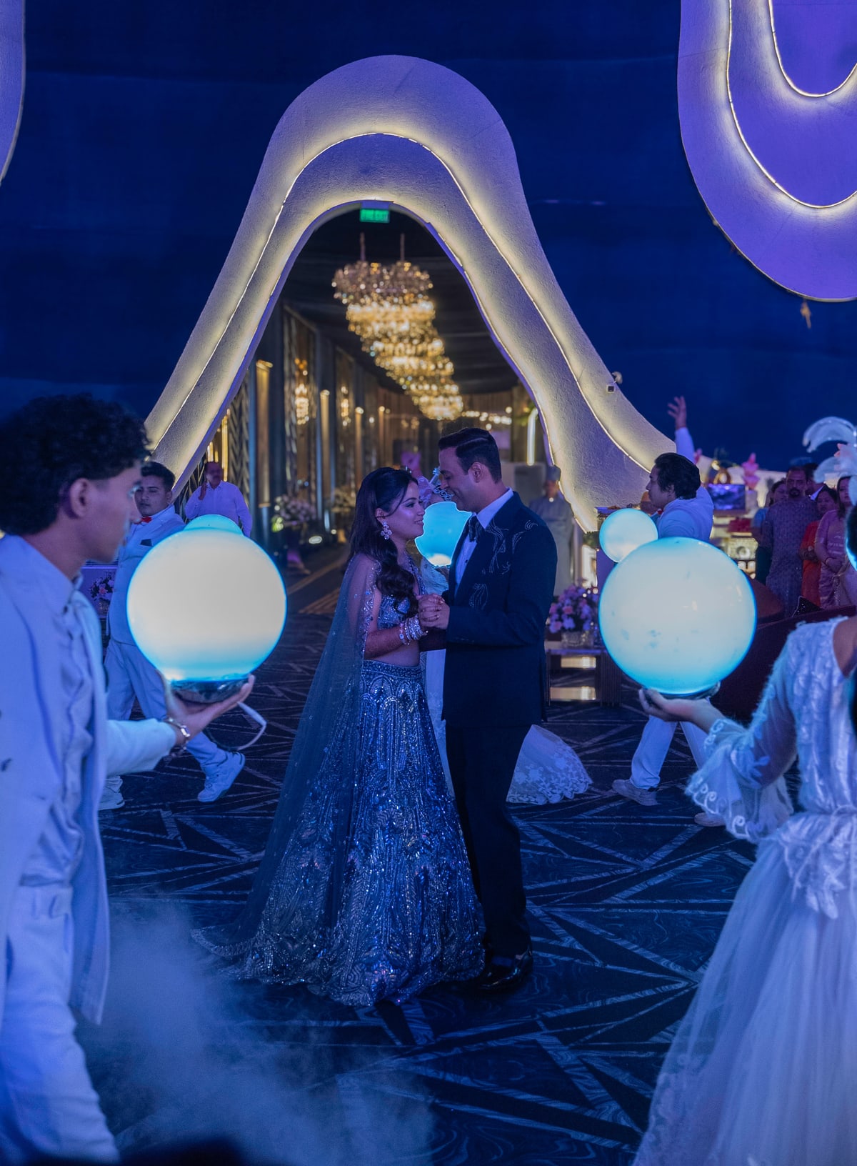 The couple walks in through a glowing archway as dancers hold illuminated orbs.