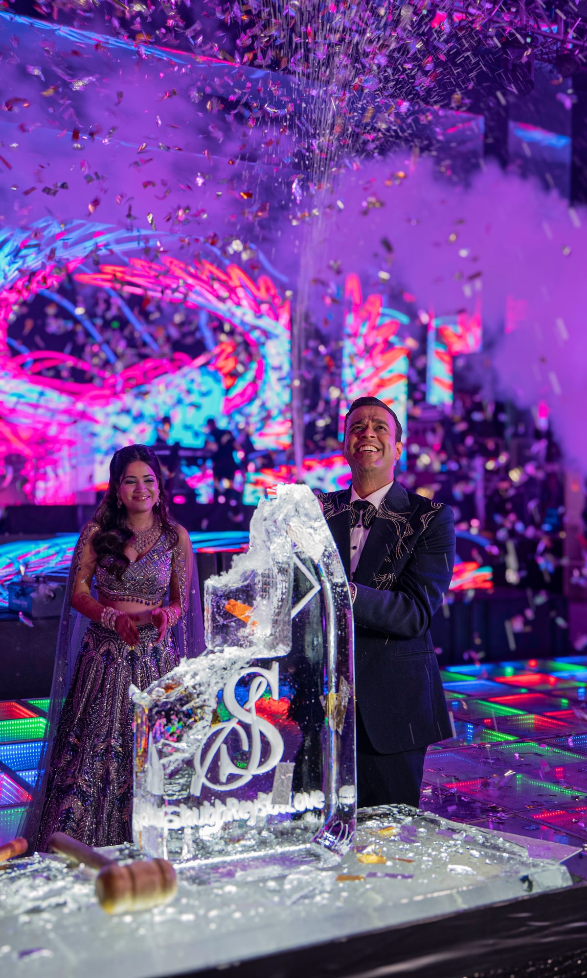A couple stands beside a monogrammed ice sculpture as confetti falls around them.