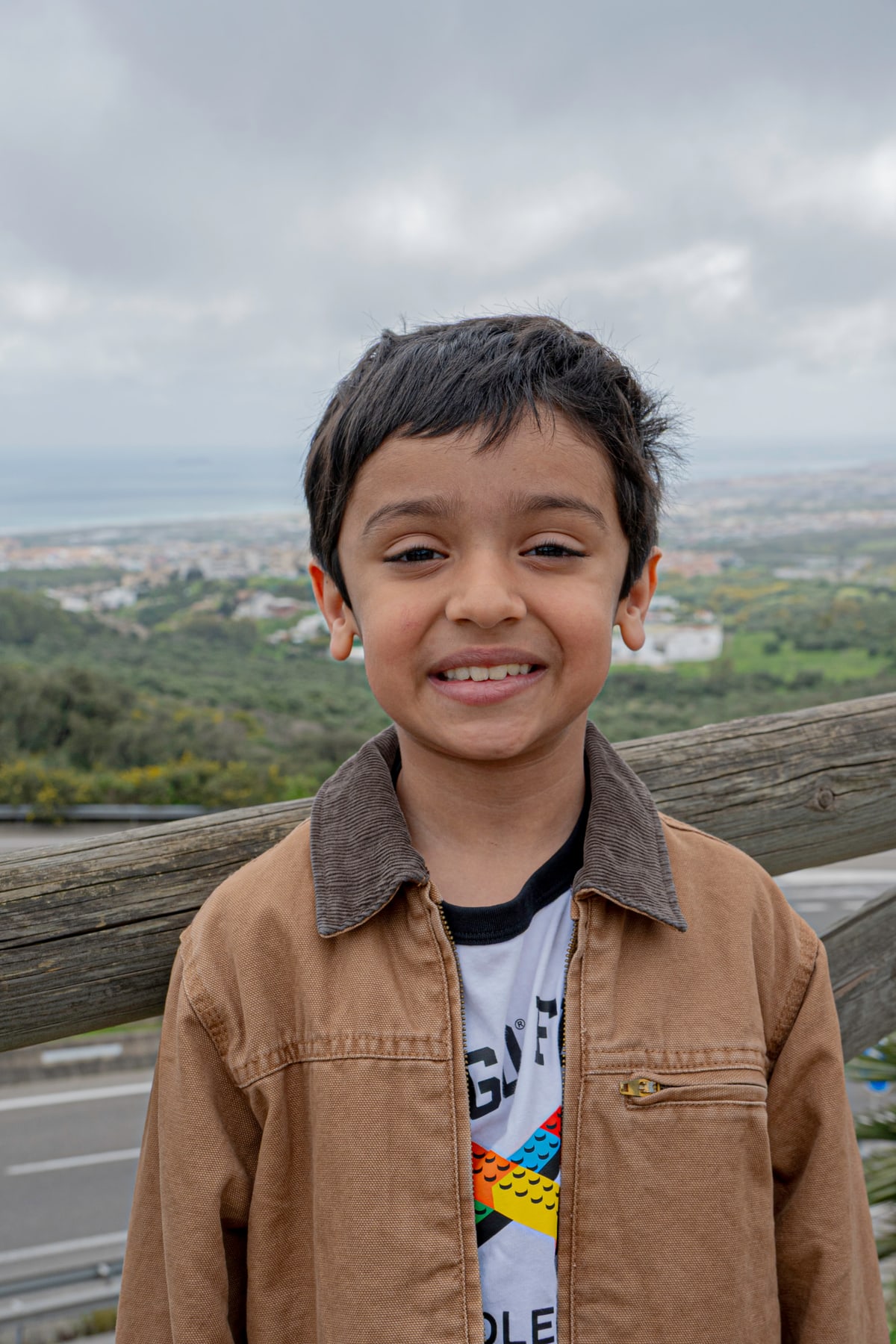 A young boy in a tan jacket grins at a hilltop overlook.
