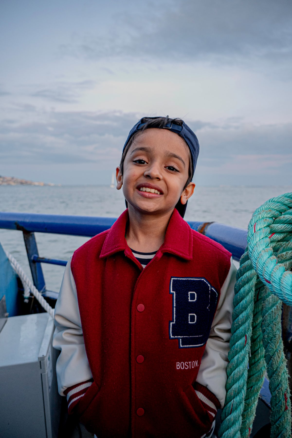 A grinning boy in a red varsity jacket stands on a boat deck.