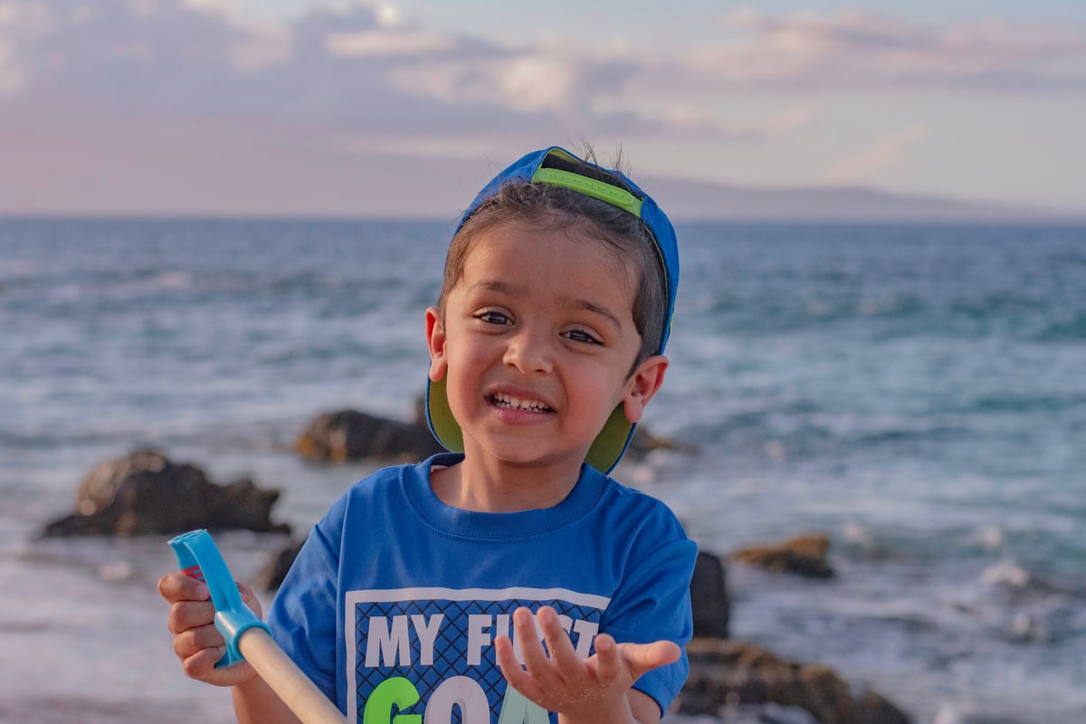 A little boy in a backwards cap grins on a rocky beach.