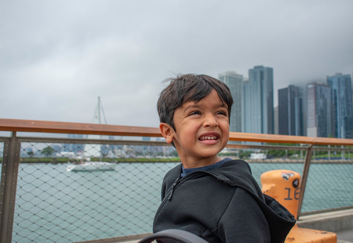 A young boy smiles at a waterfront railing with city towers behind him.