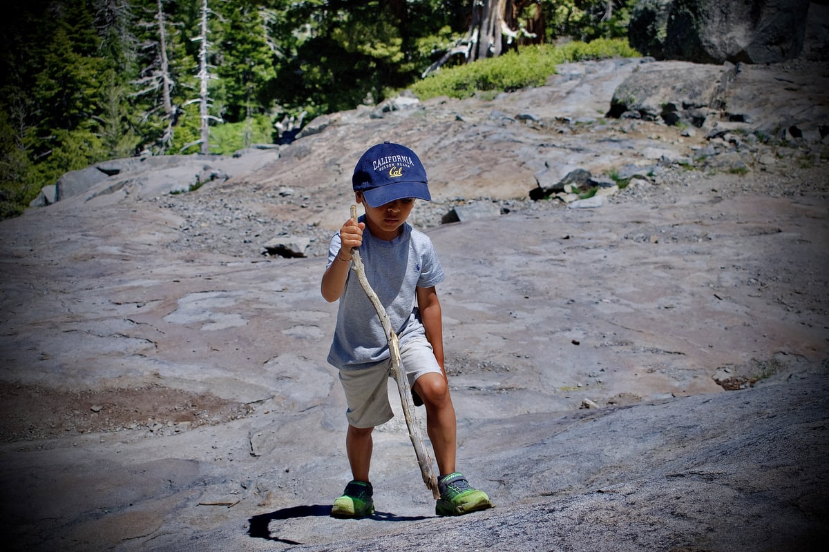 A small boy in a California cap hikes across granite with a walking stick.