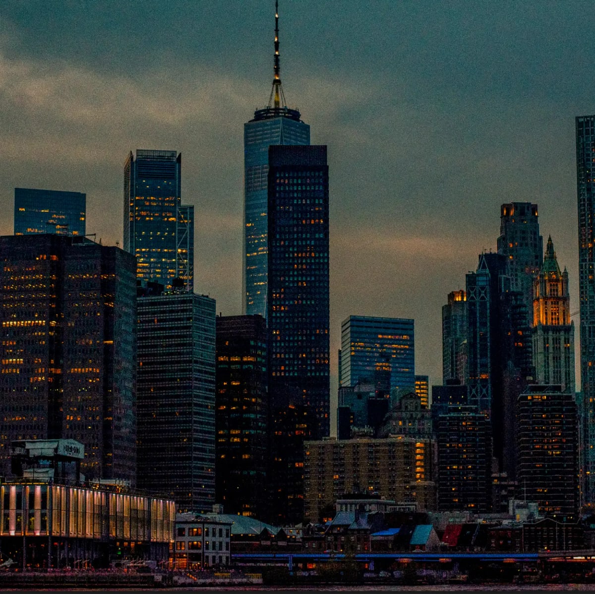 Lower Manhattan's towers glow with lit windows at dusk, One World Trade Center at the center.